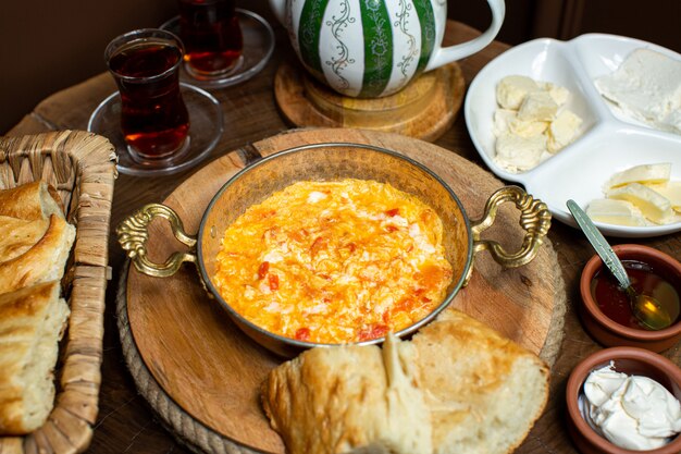 Una vista frontal de cerca huevos cocidos con tomates rojos dentro de una sartén metálica junto con té caliente y trozos de pan