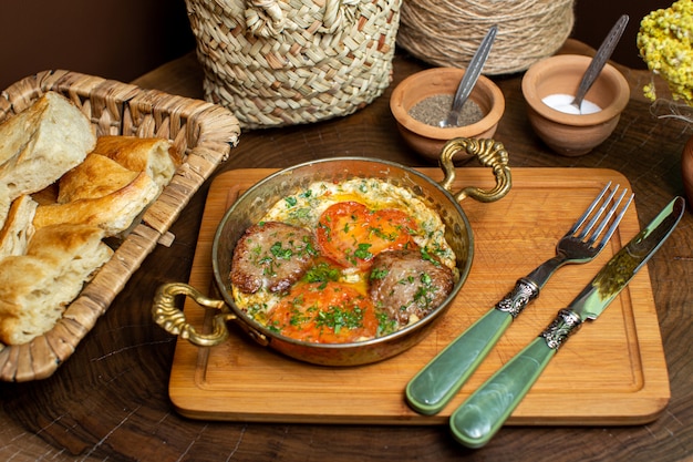 Una vista frontal de cerca huevos de comida con tomates rojos y rebanadas de carne junto con cubiertos y rebanadas de pan