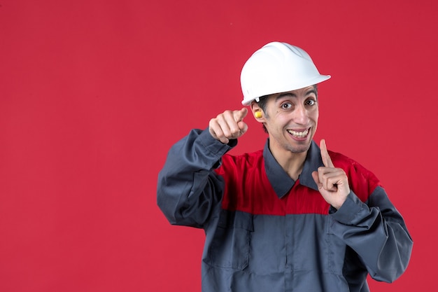Foto gratuita vista frontal cercana del confuso joven trabajador en uniforme con casco y tapones para los oídos apuntando algo en la pared roja aislada