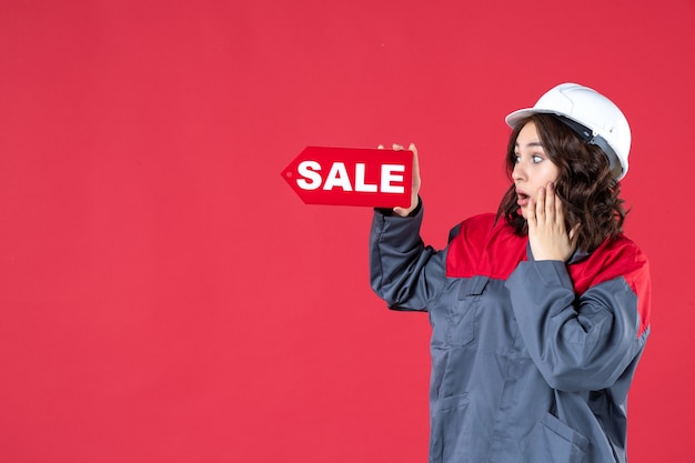 Foto gratuita vista frontal cercana de la conmocionada trabajadora en uniforme con casco y señalando el cartel de venta en pared roja aislada