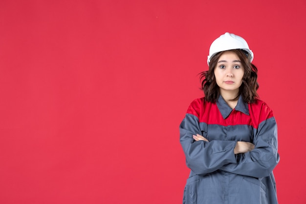 Vista frontal cercana del constructor femenino sorprendido en uniforme con casco cruzando los brazos en la pared roja aislada