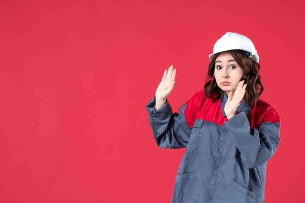 Vista frontal cercana del constructor femenino sorprendido en uniforme con casco en pared roja aislada