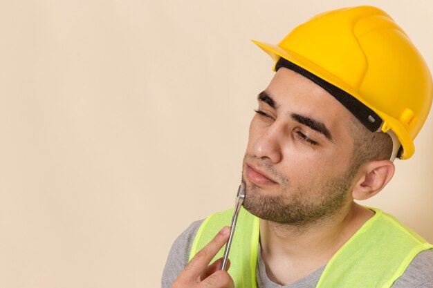 Vista frontal cercana constructor masculino en casco amarillo posando con herramienta plateada en escritorio ligero