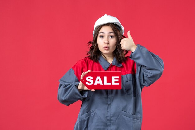 Vista frontal cercana de curiosa trabajadora en uniforme con casco mostrando el icono de venta y haciendo gesto de ok en la pared roja aislada