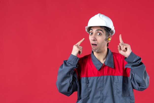 Vista frontal cercana del curioso joven trabajador en uniforme con casco y tapones para los oídos apuntando hacia arriba en la pared roja aislada