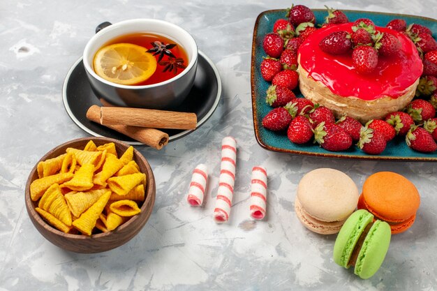 Vista frontal cercana delicioso pastel de fresa con fresas rojas frescas taza de té y macarons sobre superficie blanca