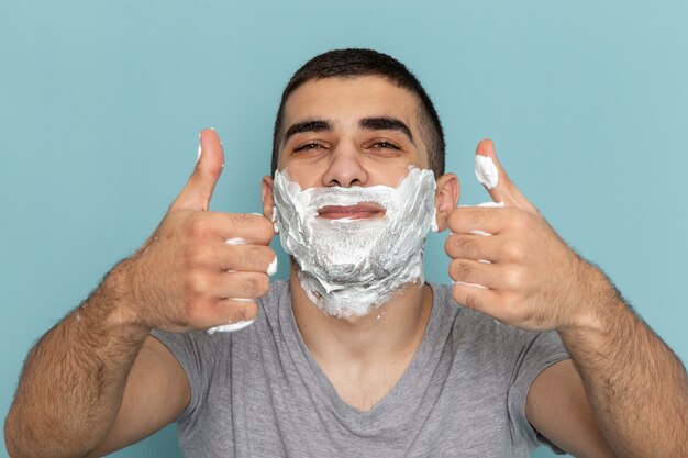 Vista frontal cercana hombre joven en camiseta gris que cubre su rostro con espuma blanca para afeitarse en la pared azul hielo espuma de barba afeitado masculino