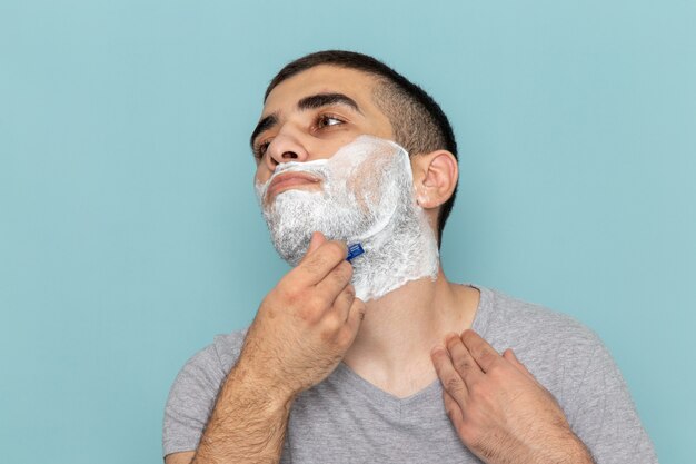 Vista frontal cercana joven en camiseta gris afeitándose la barba con navaja en la pared azul hielo barba afeitado macho