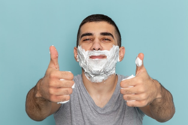 Vista frontal cercana joven en camiseta gris que cubre su rostro con espuma blanca para afeitarse en el escritorio azul hielo barba espuma afeitarse