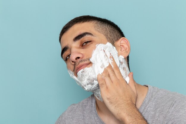 Vista frontal cercana joven en camiseta gris que cubre su rostro con espuma blanca para afeitarse en la pared azul hielo barba afeitarse con maquinilla de afeitar