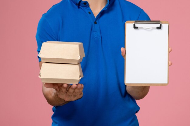 Vista frontal cercana joven mensajero en capa uniforme azul sosteniendo pequeños paquetes de comida de entrega con bloc de notas en la pared rosa claro, entrega de servicio uniforme