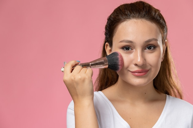 Vista frontal cercana joven mujer atractiva en camiseta blanca haciendo un maquillaje con una leve sonrisa en el fondo rosa