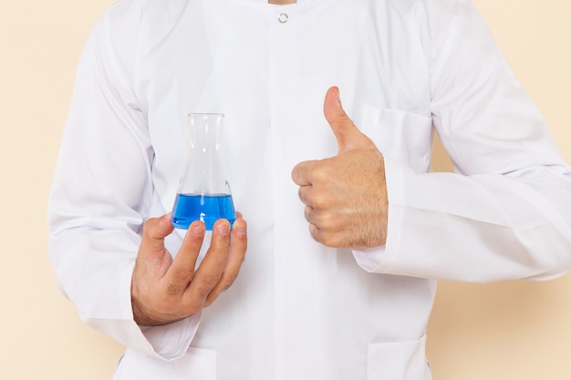 Vista frontal cercana joven químico en traje blanco especial sosteniendo un pequeño matraz con solución azul en la pared crema experimento científico química científica