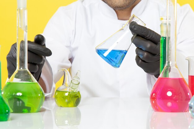 Vista frontal cercana joven químico en traje blanco frente a la mesa con soluciones coloreadas que trabajan con soluciones en el escritorio amarillo trabajo de ciencia química de laboratorio