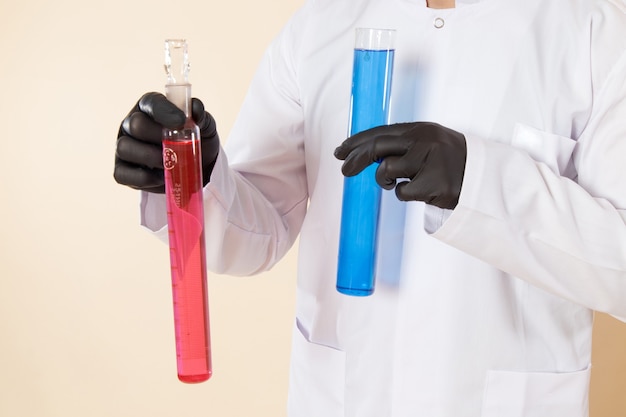 Vista frontal cercana joven químico en traje especial blanco sosteniendo pequeños frascos con soluciones coloreadas en la pared crema experimento de laboratorio de ciencia química