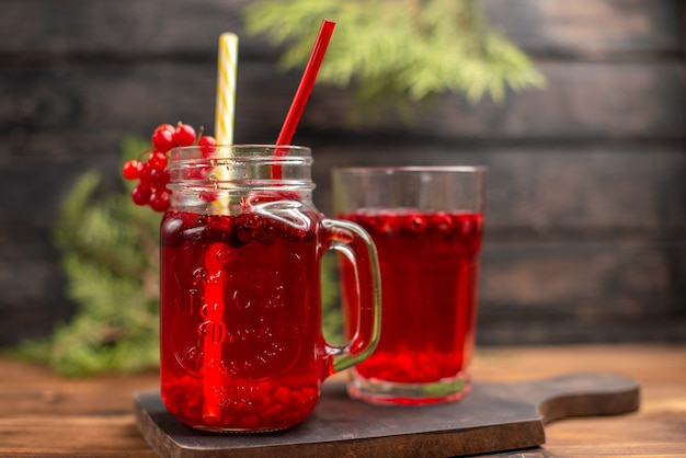 Vista frontal cercana de jugo de grosella fresca en un vaso y una taza servida con tubo sobre una tabla de cortar de madera sobre una mesa marrón