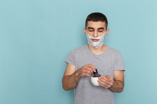 Vista frontal cercana macho joven en camiseta gris con espuma blanca en su rostro en la pared azul hielo espuma de barba macho de afeitar