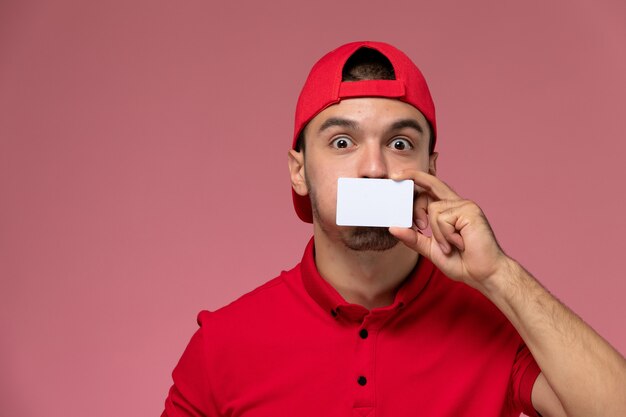 Vista frontal cercana mensajero masculino joven en gorra uniforme roja con tarjeta blanca en la pared rosa