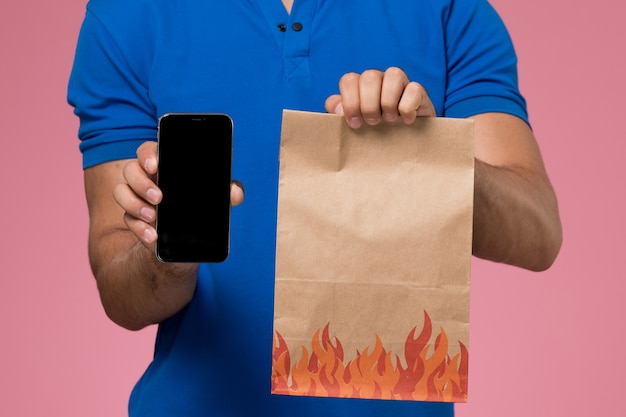 Vista frontal cercana mensajero masculino en uniforme azul con paquete de alimentos y teléfono en la entrega de trabajo de servicio uniforme rosa