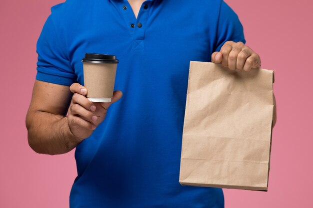 Vista frontal cercana mensajero masculino en uniforme azul con paquete de comida y taza de café en la entrega de trabajo de servicio uniforme rosa