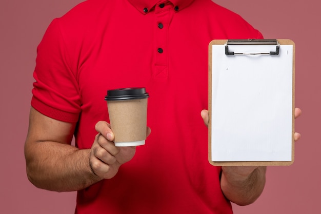 Vista frontal cercana mensajero masculino en uniforme rojo sosteniendo la taza de café de entrega y el bloc de notas en el trabajo masculino de trabajador de pared rosa