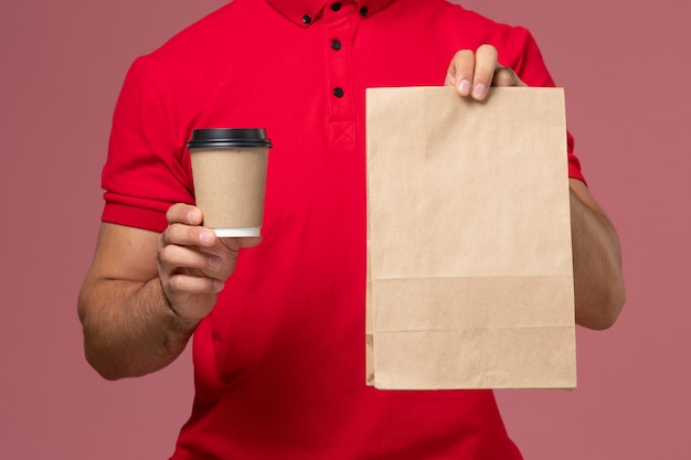 Vista frontal cercana mensajero masculino en uniforme rojo con taza de café de entrega y paquete de comida en la pared rosa servicio trabajador de entrega uniforme trabajo masculino