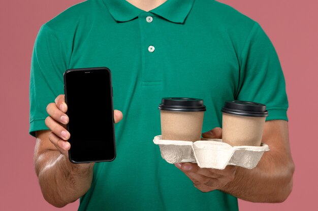 Vista frontal cercana mensajero masculino en uniforme verde sosteniendo tazas de café de entrega marrón y teléfono en el trabajo de escritorio rosa