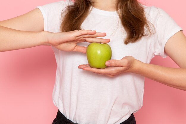 Vista frontal cercana mujer joven en camiseta blanca con manzana verde en la pared rosa