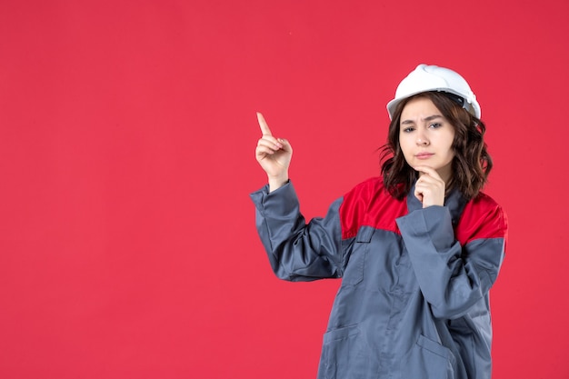 Vista frontal cercana del pensamiento constructor femenino en uniforme con casco y apuntando hacia arriba en la pared roja aislada