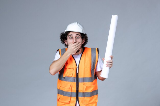 Vista frontal cercana del trabajador de la construcción confundido en chaleco de advertencia con casco de seguridad y sosteniendo el espacio en blanco en la pared gris