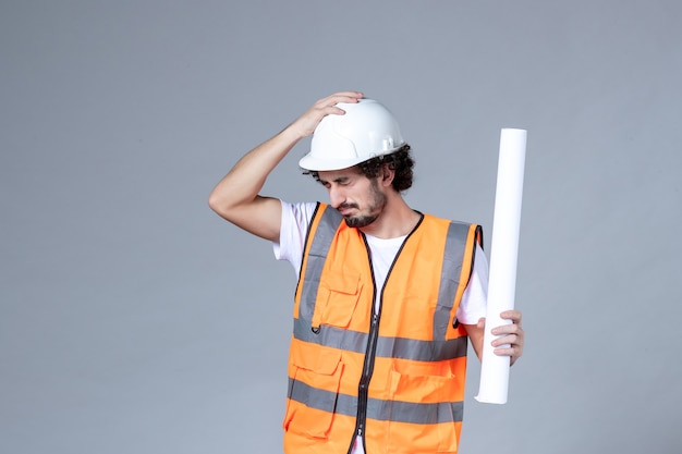 Vista frontal cercana del trabajador de la construcción masculino cansado en chaleco de advertencia con casco de seguridad y sosteniendo el espacio en blanco en la pared gris