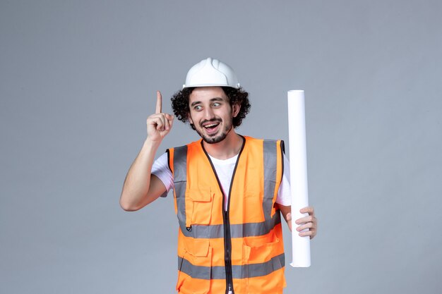 Vista frontal cercana del trabajador de la construcción masculino sonriente en chaleco de advertencia con casco de seguridad y mostrando en blanco apuntando hacia arriba en la pared gris