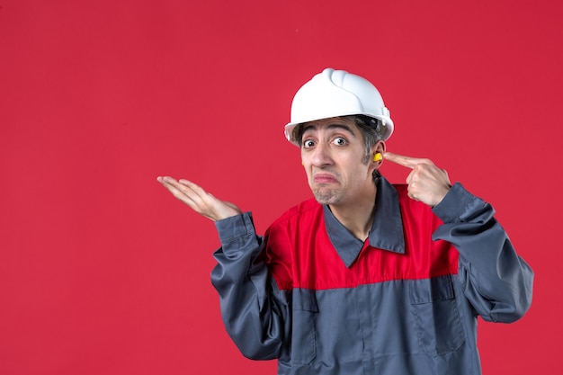 Foto gratuita vista frontal cercana del trabajador joven confundido en uniforme con casco y tapones para los oídos apuntando en la pared roja aislada