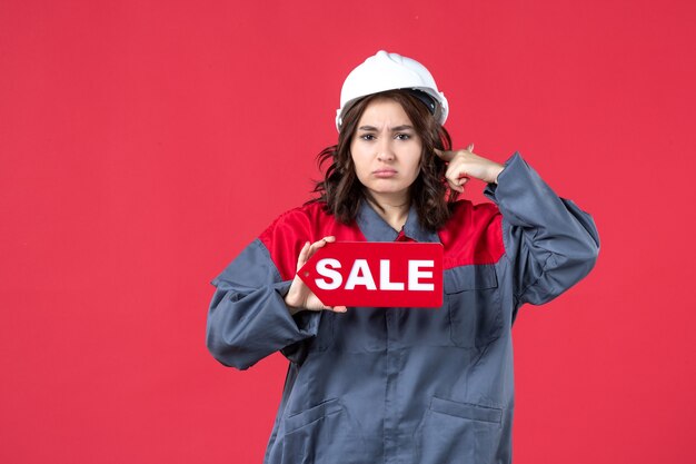 Vista frontal cercana de la trabajadora agresiva en uniforme con casco mostrando el icono de venta en la pared roja aislada