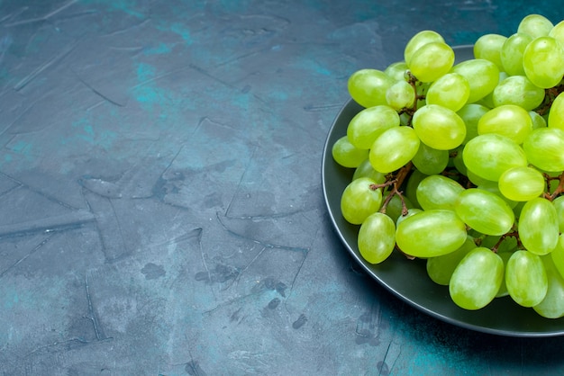 Vista frontal cercana uvas verdes frescas frutas jugosas y suaves en el escritorio de color azul claro.