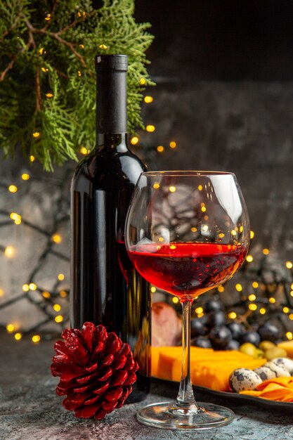 Vista frontal cercana de vino tinto seco en un vaso y en una botella junto al aperitivo y el cono de coníferas sobre fondo gris