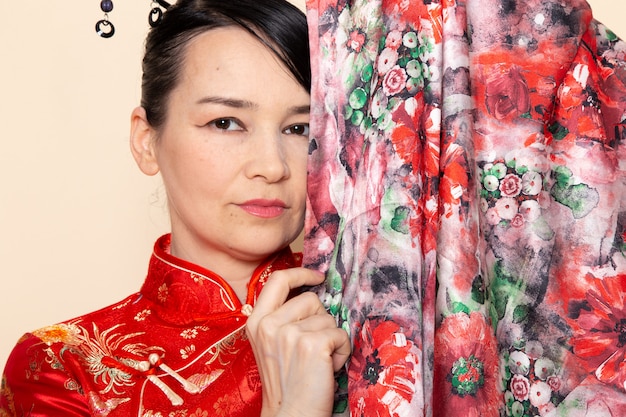 Una vista frontal cerrada exquisita geisha japonesa en vestido tradicional rojo japonés posando con tejido de flores diseñado elegante sonriendo en la ceremonia de fondo crema Japón