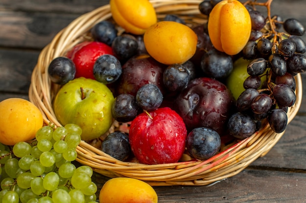 Vista frontal de la cesta con frutas suaves y amargas, como uvas, albaricoques, ciruelas en el escritorio rústico marrón fruta