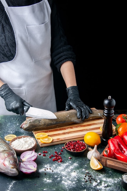 Vista frontal del chef en delantal para picar pescado crudo en tablero de madera molinillo de pimienta tazón de harina semillas de granada en un tazón en la mesa de la cocina