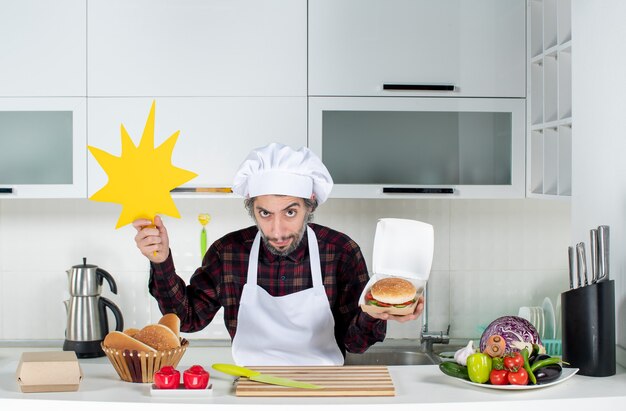 Vista frontal del chef escéptico sosteniendo un cartel de boom crunch y una hamburguesa en la cocina moderna