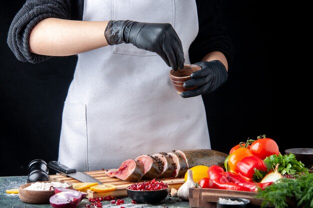 Vista frontal del chef espolvoreado con pimienta negra en rodajas de pescado crudo en la tabla de cortar verduras en la tabla de servir de madera en la mesa de la cocina