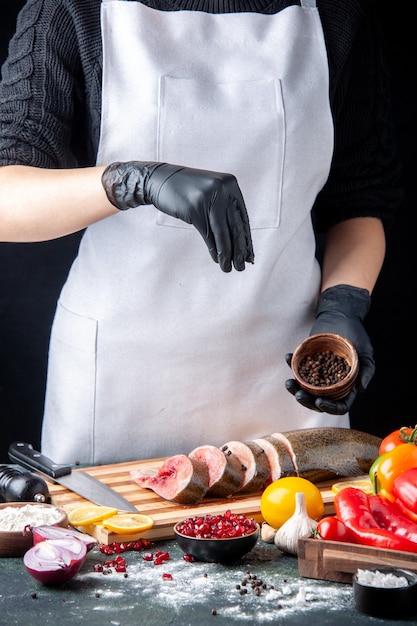 Vista frontal del chef espolvoreado con pimienta negra en rodajas de pescado crudo en la tabla de cortar verduras en la tabla de servir de madera en la mesa de la cocina