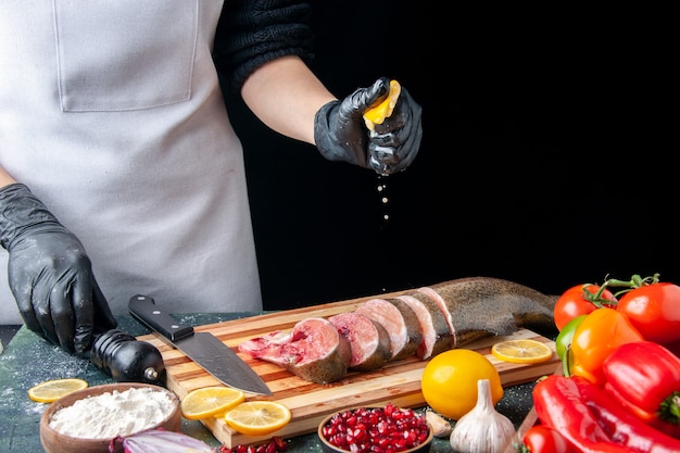 Vista frontal del chef exprimiendo limón en rebanadas de pescado crudo cuchillo en tabla de cortar verduras en tabla de servir de madera en la mesa de la cocina