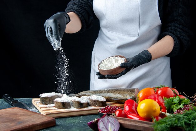 Vista frontal de la chef femenina que cubre rebanadas de pescado crudo con harina de verduras frescas en tablero de madera cuchillo de tazón de harina en la mesa de la cocina