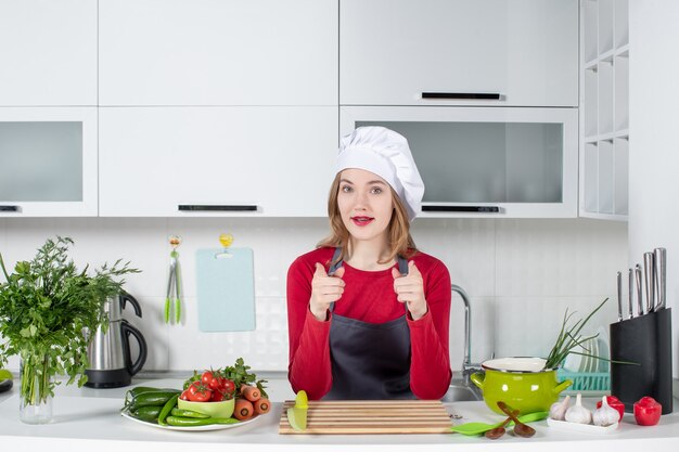 Vista frontal chef femenina en uniforme de pie detrás de la mesa de la cocina apuntando al frente