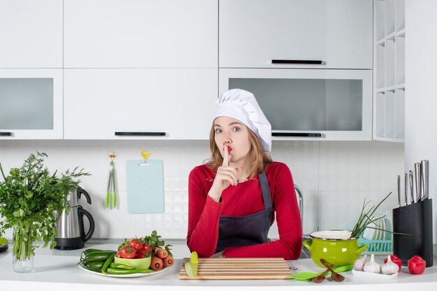 Vista frontal chef femenina en uniforme de pie detrás de la mesa de la cocina haciendo señal de silencio