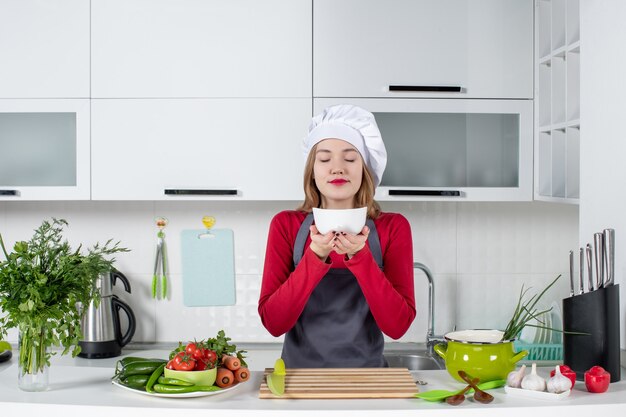 Vista frontal chef femenina en uniforme de pie detrás de la mesa de la cocina sosteniendo un tazón oliendo algo