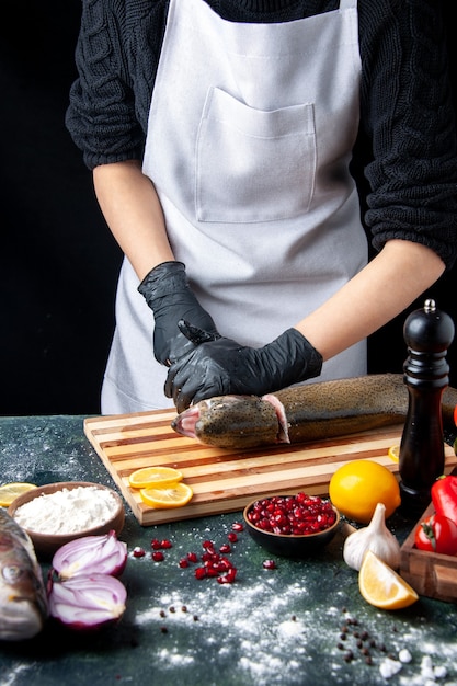 Vista frontal del chef con guantes negros cortando la cabeza de pescado en una tabla de cortar, molinillo de pimienta, tazón de harina, semillas de granada en un tazón en la mesa de la cocina