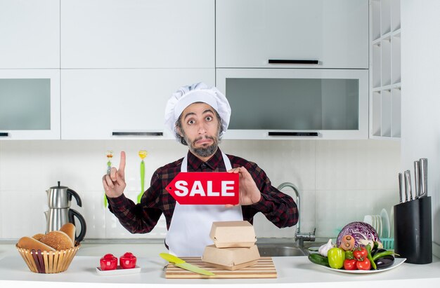 Vista frontal del chef masculino apuntando al techo sosteniendo cartel de venta en la cocina