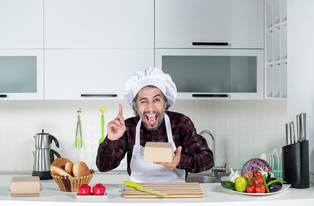 Vista frontal del chef masculino apuntando con el dedo hacia arriba sosteniendo la caja en la cocina